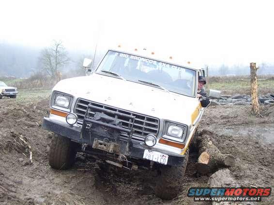 1978 Ford Bronco stuck in mud with a log under its tire. Another vehicle is visible in the background