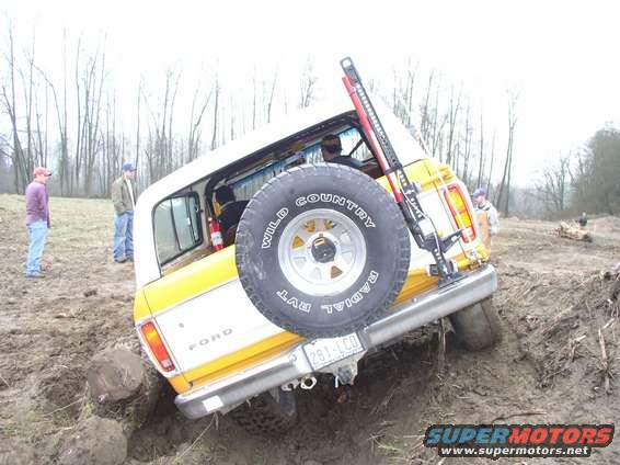 Yellow and white 1978 Ford Bronco tilted in mud, spare tire mounted on rear door, high-lift jack attached