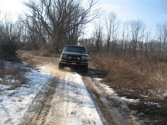 1996 Ford Bronco driving on a muddy, snowy road with trees in the background