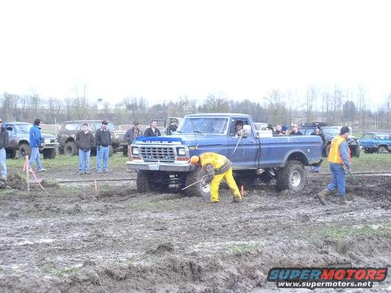 Front view of a blue 1979 Ford F-Series pickup truck in mud, with a person in yellow working on the front tire