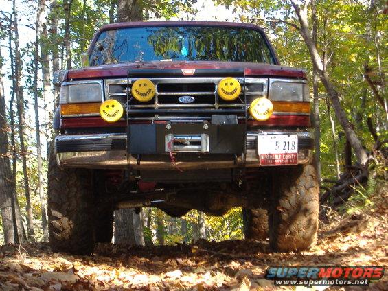 Red Ford truck with large tires and a winch, viewed from the front in a wooded area