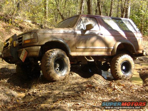 Muddy brown 1996 Ford Bronco off-roading through mud and trees