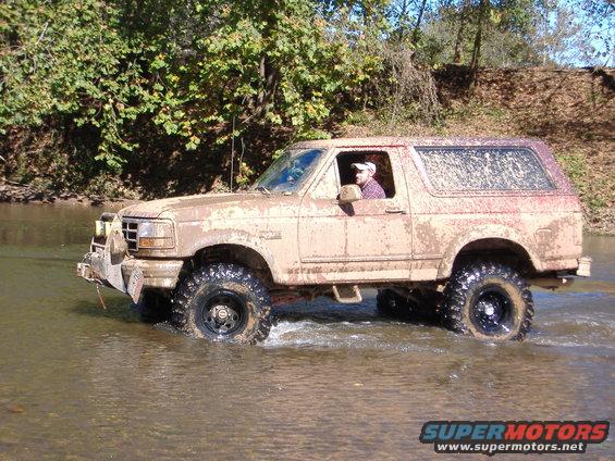Muddy SUV driving through a shallow river with trees in the background