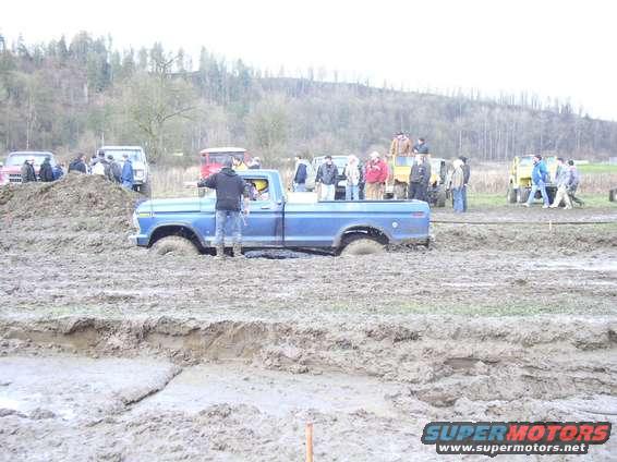 Blue pickup truck stuck in mud pit with people watching