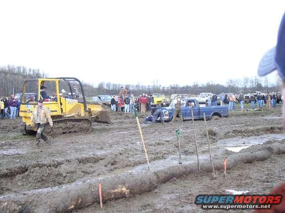 Yellow bulldozer and blue pickup truck stuck in mud, with spectators watching