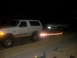 1992 Ford Bronco - A white 1992 Ford Bronco is seen from the side, towing a car on a trailer in the dark.