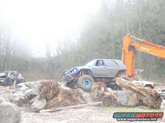 Modified off-road SUV climbing over logs and rocks, with an excavator in the background