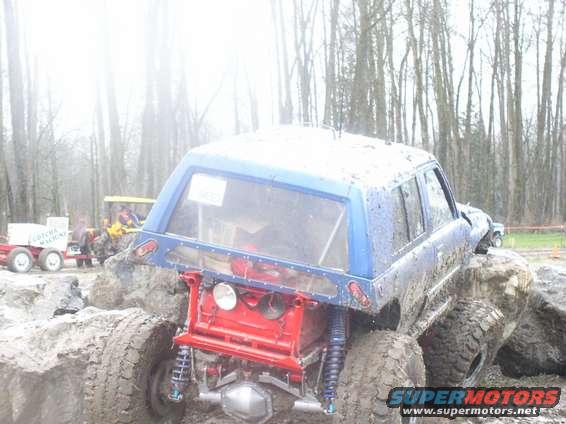 Rear view of blue off-road SUV with exposed red frame and large tires stuck in mud