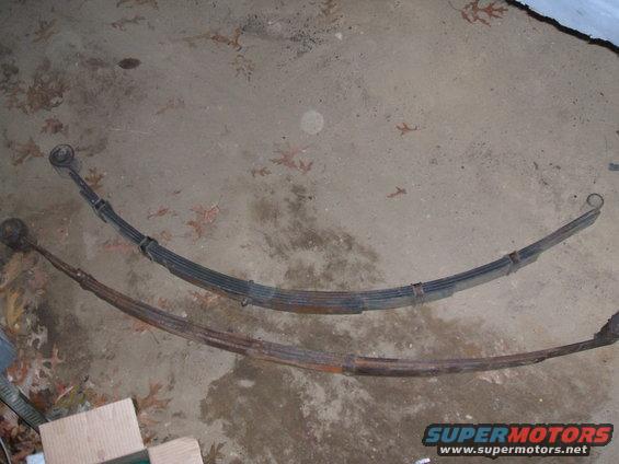 Two leaf springs on a concrete floor. One is black and appears newer, the other is rusty and older