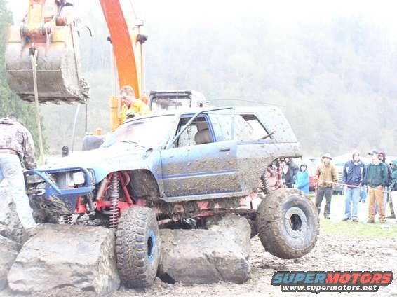 Muddy SUV with lifted suspension and large tires is being lifted by an excavator. People watch in the background