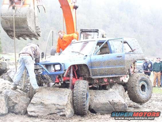 Mud-covered blue SUV with oversized tires on rocks, front bumper removed, driver's door open