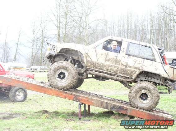 Mud-covered SUV with large tires on a wooden ramp, driver visible