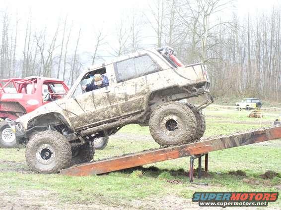 Mud-covered off-road SUV on a wooden ramp, with a red truck behind it