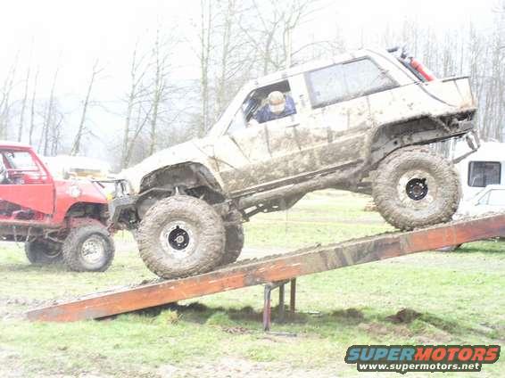 Muddy off-road vehicle climbing a ramp, with a red truck nearby