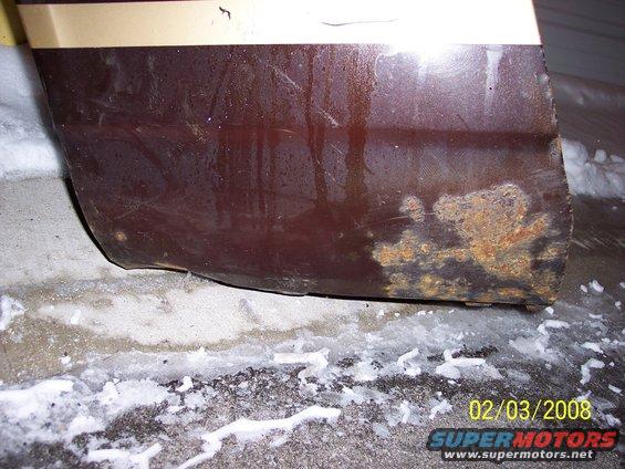 close-up of a brown vehicle's lower rear quarter panel with a tan stripe above, showing significant rust damage near the bottom edge