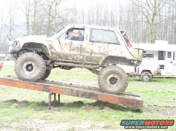 Muddy off-road vehicle with large tires on a wooden ramp; a white trailer is parked behind it