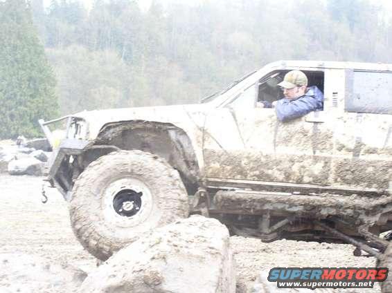 Muddy off-road vehicle with large tires, driver wearing a cap and jacket, navigating rough terrain