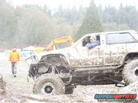 Mud-caked off-road SUV with exposed suspension, driver in window, parked in mud