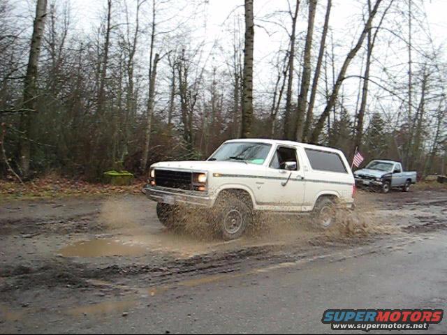 1980 Ford Bronco splashing through a muddy road with a pickup truck in the background