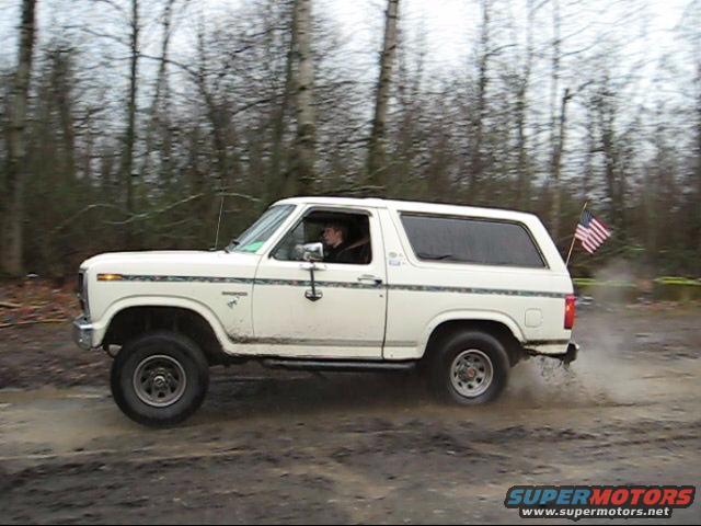 White 1980 Ford Bronco driving through mud with an American flag on the back
