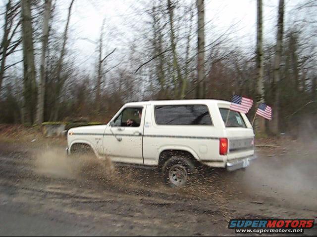 White 1980 Ford Bronco driving through mud with two American flags on the back
