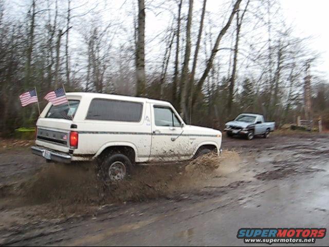 White 1980 Ford Bronco splashing through mud with American flags on rear