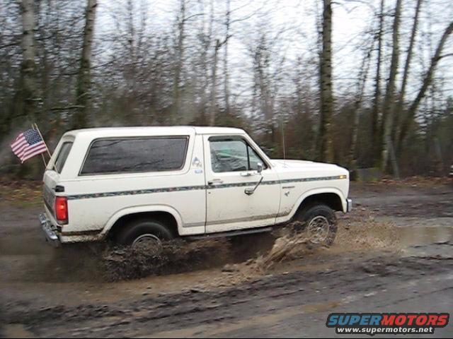 White Ford Bronco with American flag on back driving through mud puddle