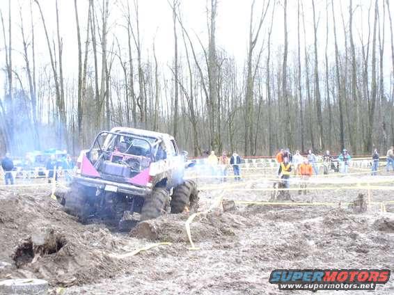 Rear view of a modified off-road truck with a roll cage stuck in mud, emitting smoke