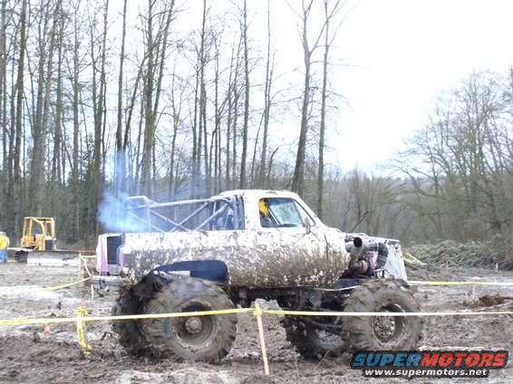 Mud-covered monster truck with large tires and exposed engine, emitting blue smoke from exhaust
