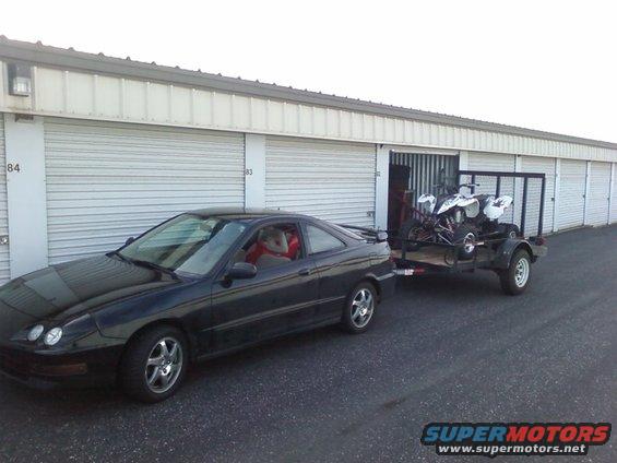 Black coupe towing a trailer with two ATVs, parked in front of storage units