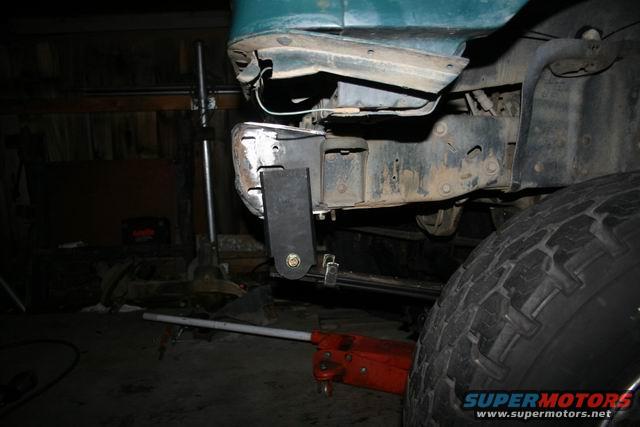 Close-up of a custom bracket being bolted to a vehicle's frame, likely for a bumper modification. A red hydraulic jack is visible underneath