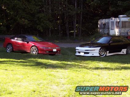 red sports coupe on left, black and white sports coupe on right, parked on grass with trees and trailer behind