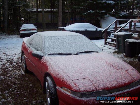 Red coupe with snow on its roof and hood, parked outside. The front end is visible, showing pop-up headlights