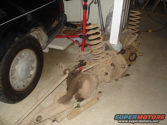 Disassembled rear axle with springs and shocks, next to a black vehicle's rear wheel well showing rust
