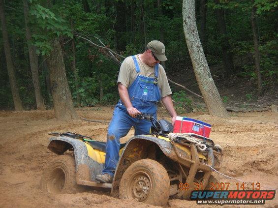 Man in overalls on a yellow ATV stuck in mud, with a cooler on the back