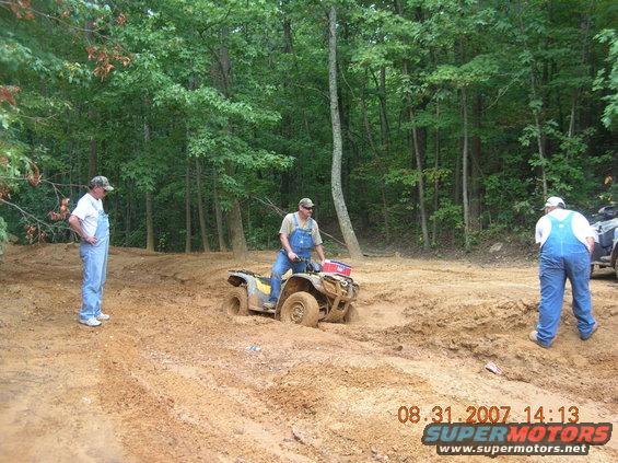 Yellow all-terrain vehicle stuck in mud with a man riding it and two men standing nearby