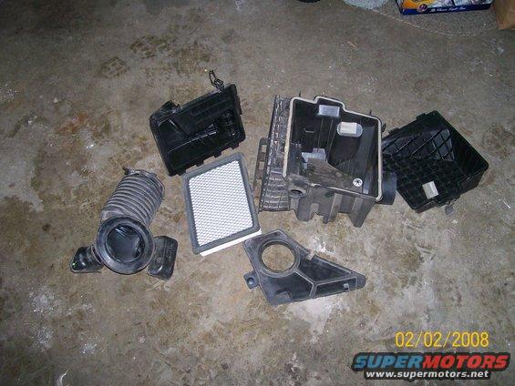 Disassembled black plastic air intake system parts and a white air filter on a concrete floor