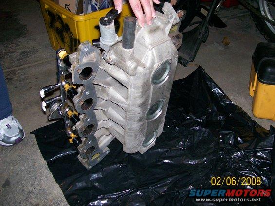 Close-up of an aluminum intake manifold with fuel injectors attached, resting on a black tarp