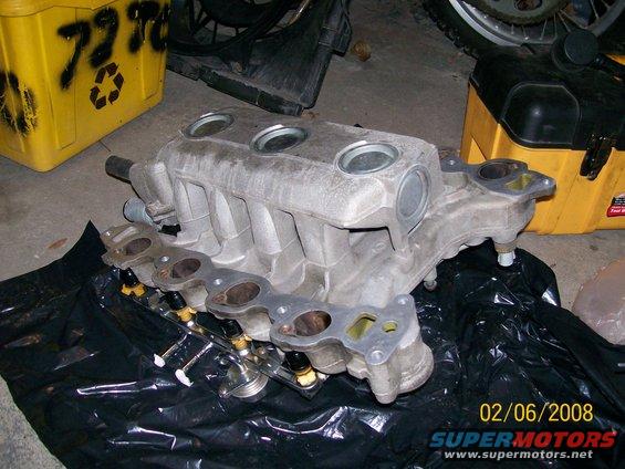 Aluminum intake manifold with fuel injectors attached, resting on black plastic