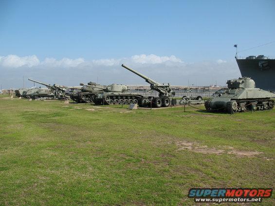 Row of tanks and artillery pieces with a large ship in the background