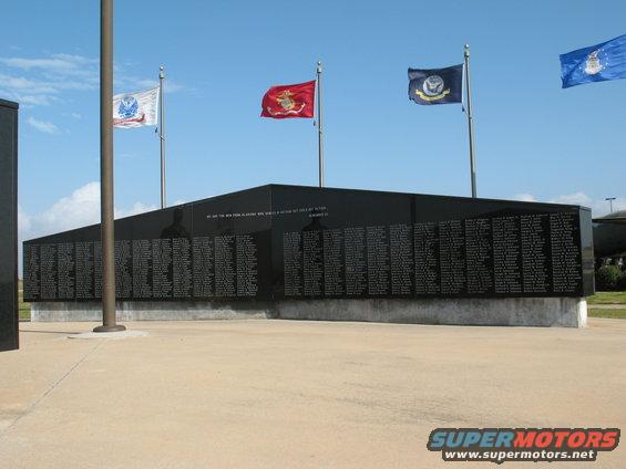Black granite war memorial wall with names inscribed, under a blue sky with flags