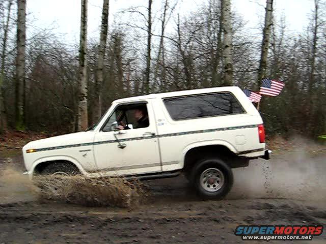 White 1980 Ford Bronco driving through mud with American flags on the back