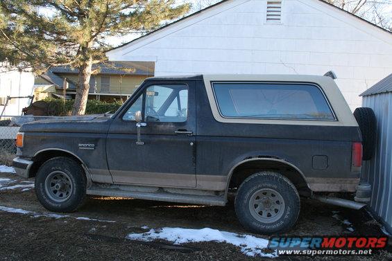 Dark gray Ford Bronco with a tan top, seen from the side. The vehicle is dirty and parked next to a metal shed