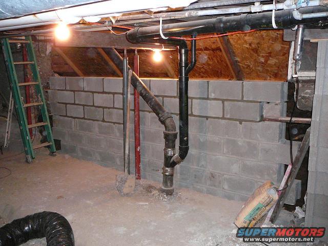 Unfinished basement wall with cinder blocks, plumbing pipes, and a ladder
