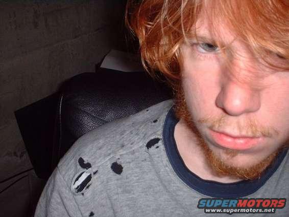 Close-up of a person with red hair and beard, wearing a torn grey t-shirt. Text 'SUPERMOTORS' visible at bottom right