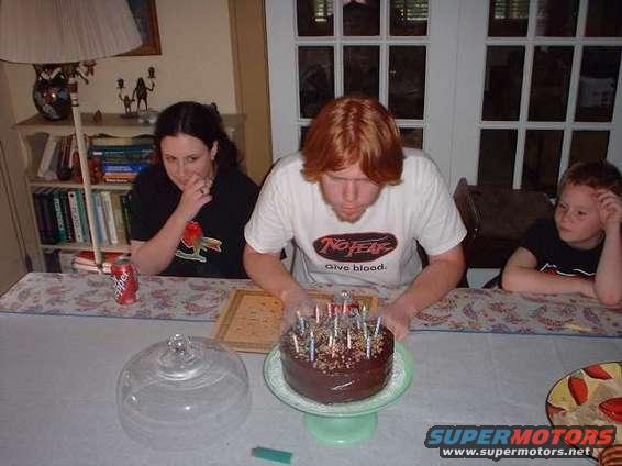Three people gathered around a table with a chocolate birthday cake and lit candles