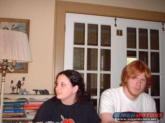 Two people sit in a room with a lamp and bookshelves
