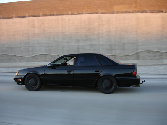 1991 Ford Taurus driving on highway, side view. Black paint, tinted windows, stock wheels