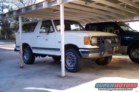 White 1989 Ford Bronco parked under a carport next to a dark pickup truck