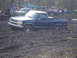 1989 Ford Bronco - A blue pickup truck is stuck deep in the mud, with spectators watching from the side and other vehicles in the background.
